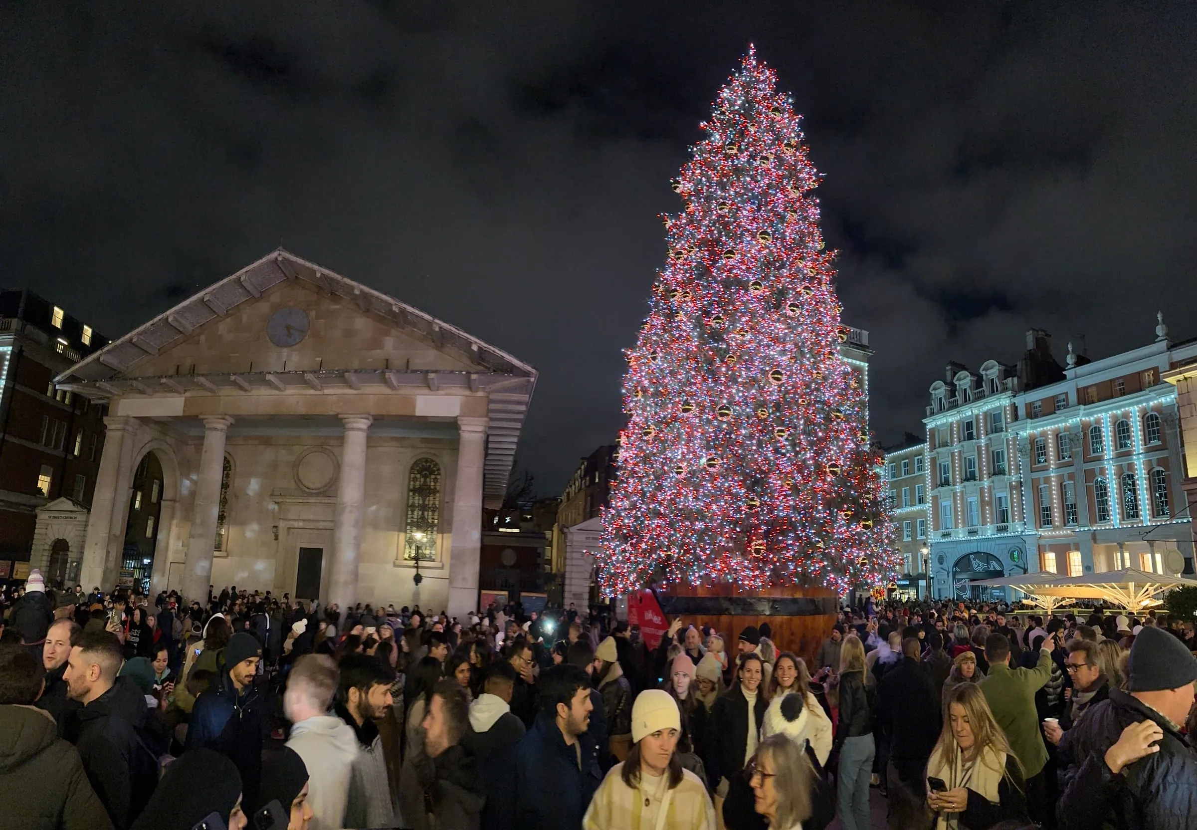 A tall Christmas tree covered in red and white lights on Covent Garden's West Piazza beside the portico of St Paul's Church, with a large crowd in front and lit building fronts on the right