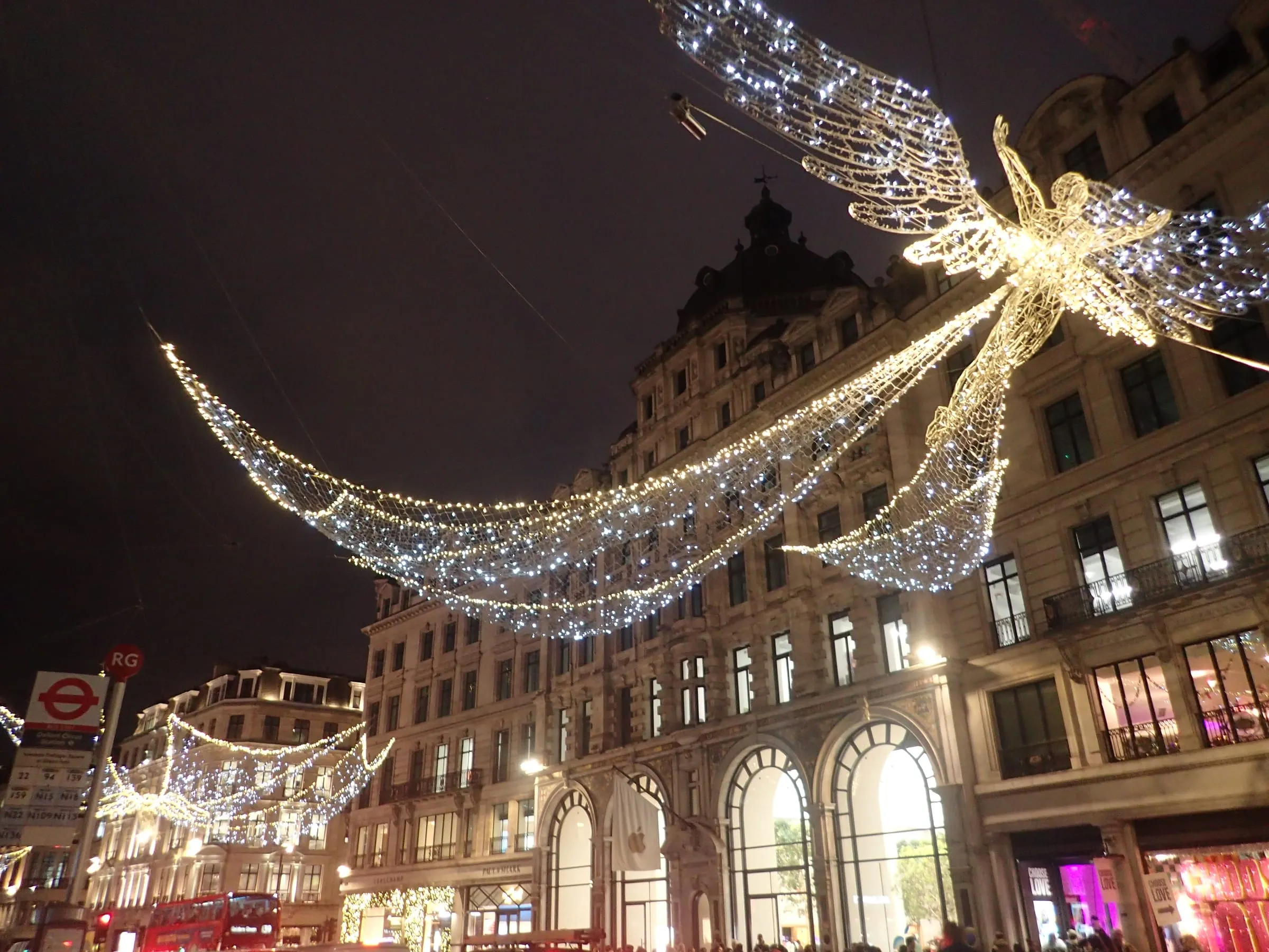 A Spirit of Christmas angel above Regent Street at night, its lit wings spanning the road between strings of white lights, with the Apple store's arched windows below