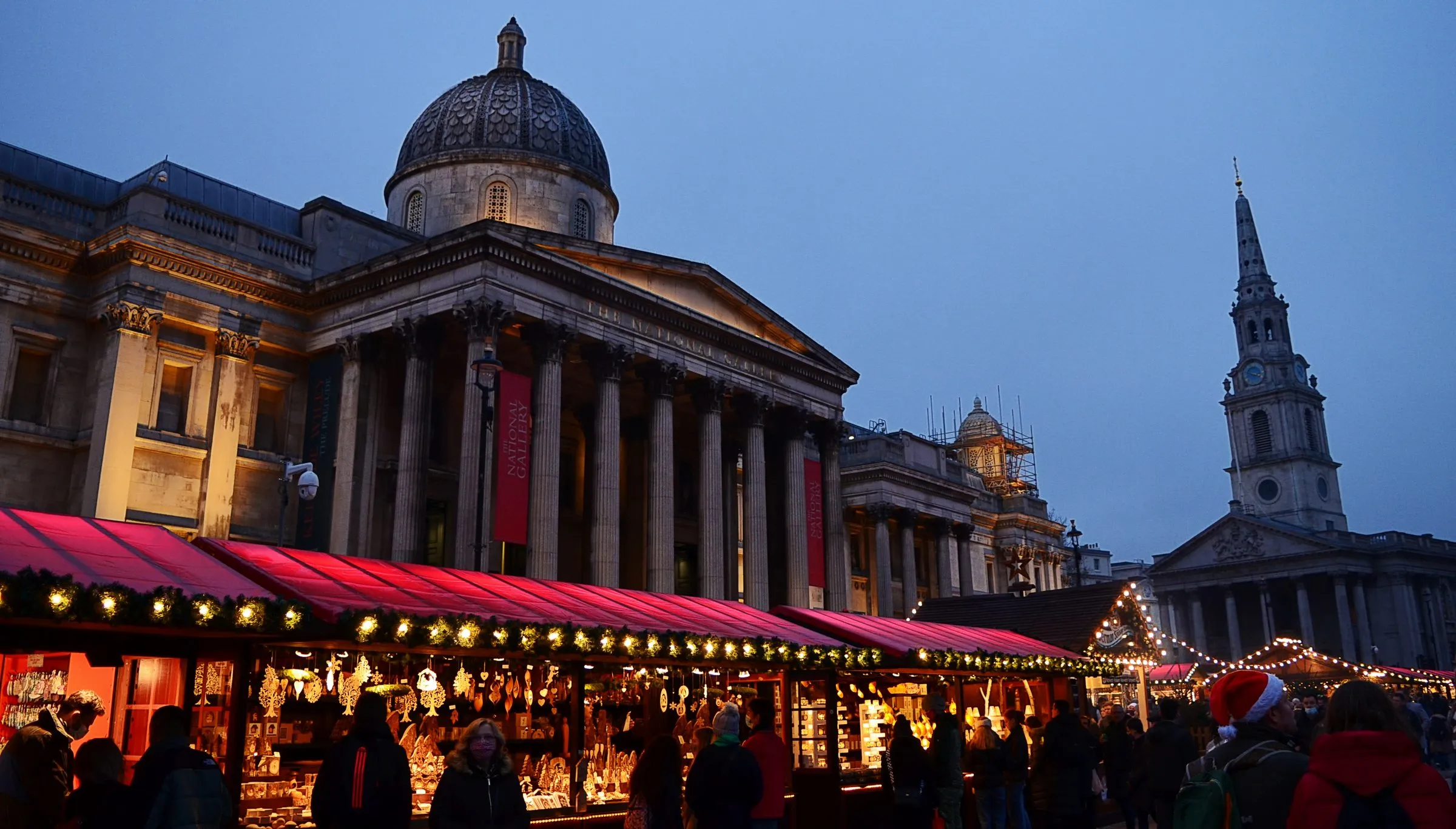 Red-roofed wooden chalets of the Trafalgar Square Christmas market lit with garlands at dusk, in front of the National Gallery's portico and dome, with the spire of St Martin-in-the-Fields on the right